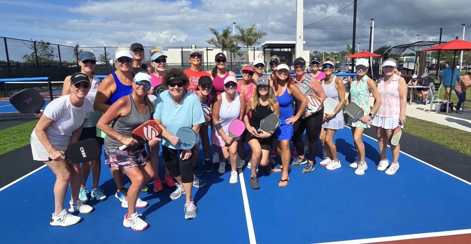 A big group of Jill Jacobs' pickleball friends on a blue court, paddles in hand